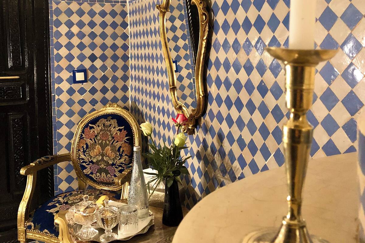 A room with a blue and white checkered tile wall, a black door, a gold-framed mirror, a table with a vase and a candle holder, and a blue and gold ornate chair.