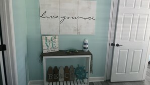 A hallway with a light blue wall, a white door, a white console table with a sign saying "LOVE you to the beach and back," a decorative ship wheel, and a wall art piece with the text "love you more." - Hidden in Paradise/ Pet friendly/ private house! (Gulf Shores)