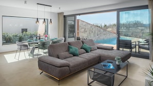 A modern living room with a large gray sectional sofa, a dining area with a glass table, and a view of a swimming pool. - Nice home in Stancijeta (Stancijeta)