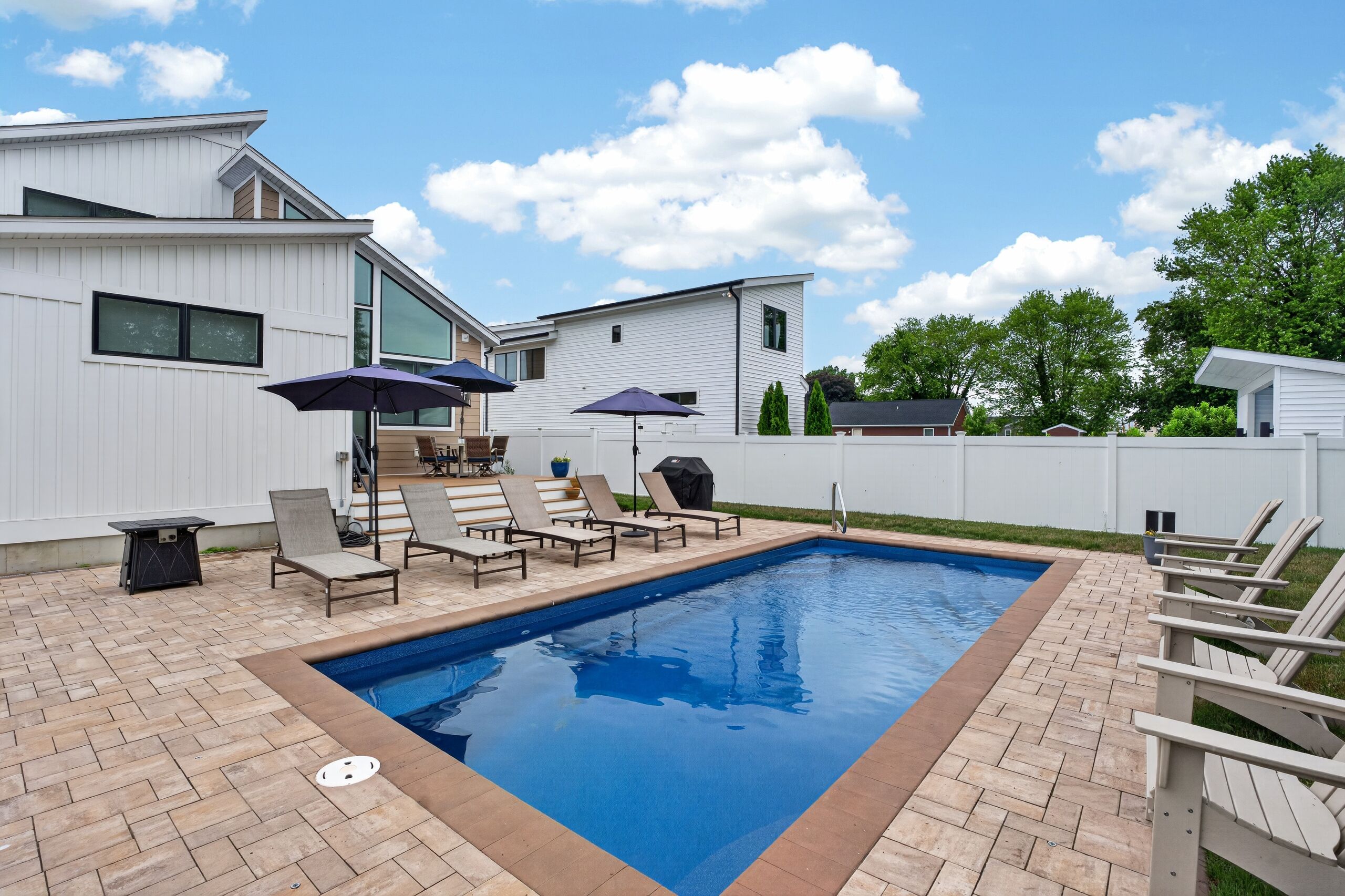 A swimming pool with lounge chairs and umbrellas, surrounded by a brick patio and a white fence.