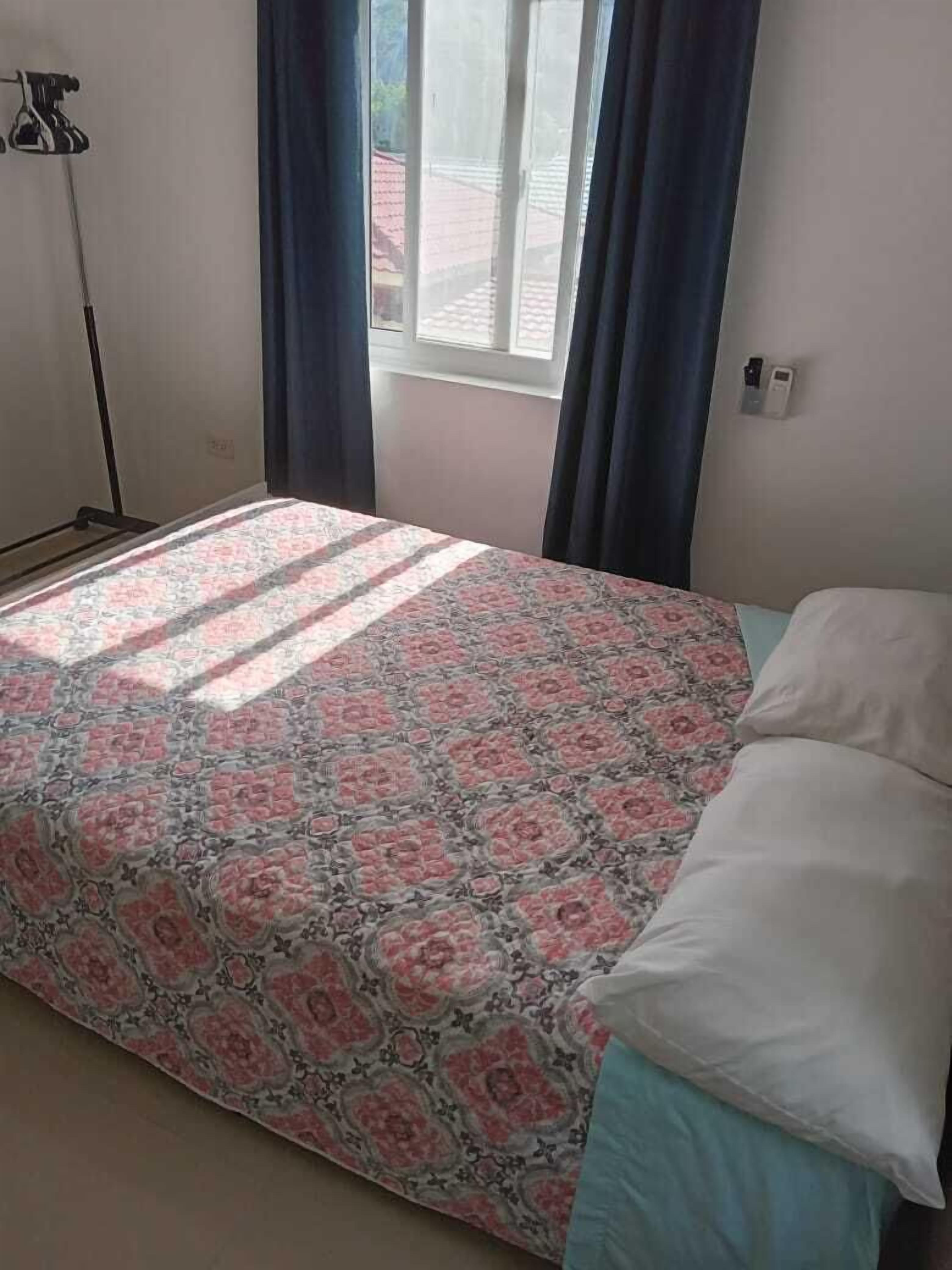 Un dormitorio con una colcha estampada, una cama individual con almohadas blancas, una lámpara de pie y un ventanal con cortinas.