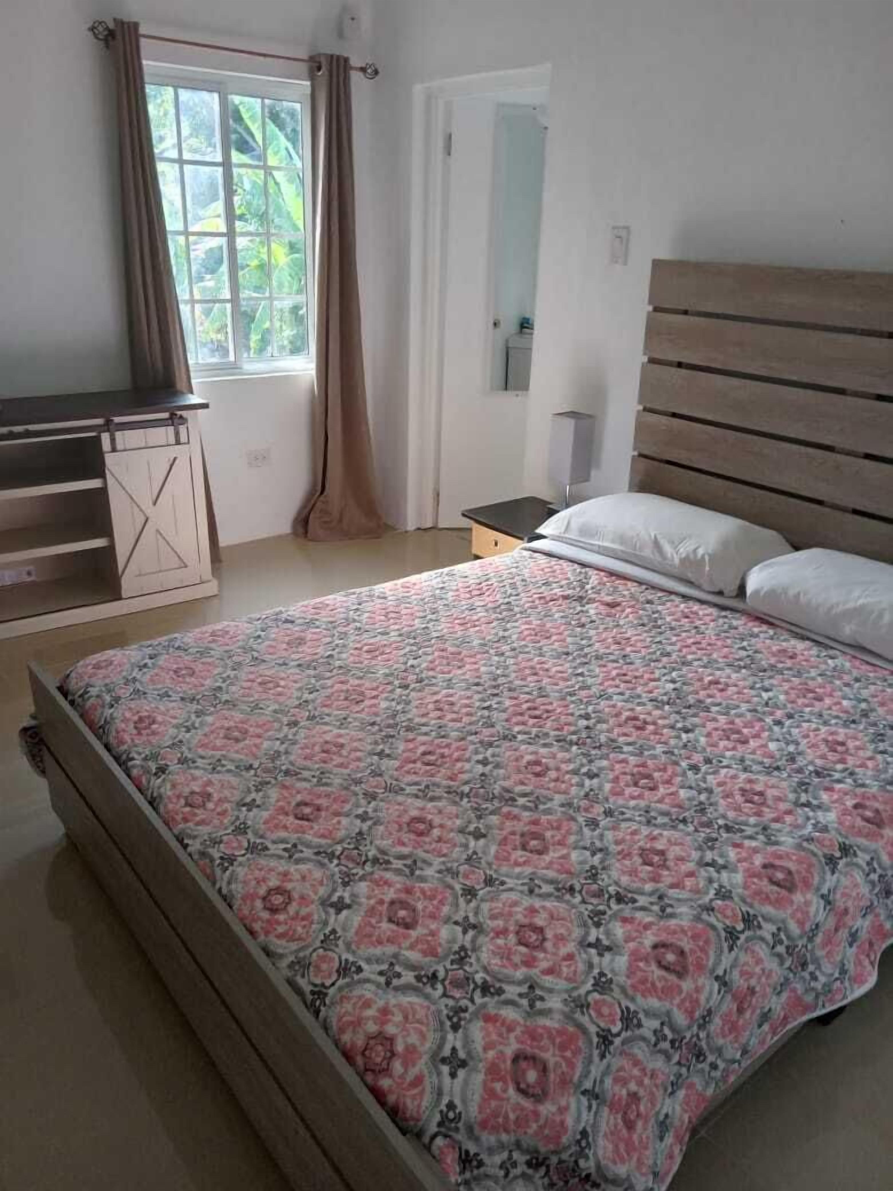 Un dormitorio con una cama de madera, una mesita de noche, una ventana con cortinas y una puerta.
