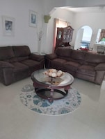 Uma sala de estar com dois sofás marrons, uma mesa de centro de vidro e um tapete floral, além de uma área de jantar com mesa posta para dois.
