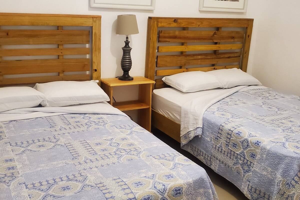 A room with two wooden beds, a nightstand with a lamp, and two framed pictures on the wall.
