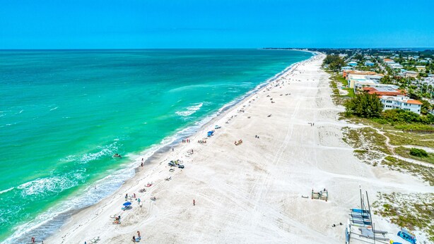 Une plage aux eaux turquoise et limpides, un rivage sablonneux et une rangée de bâtiments avec des toits rouges qui s’étendent sur le côté droit.