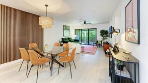 Un salon moderne avec une table à manger en verre, des chaises orange, un mur d’accent en bois et une grande fenêtre donnant sur un paysage verdoyant.