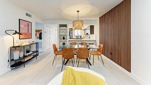 Un espace repas moderne avec une table en verre et des chaises marron, une console noire agrémentée d’objets décoratifs et un mur en lambris de bois sur le côté droit.