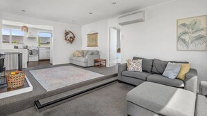 Una sala de estar moderna con un sofá gris, una mesa de centro y un tapete estampado. Se ve una cocina al fondo con armarios blancos y una lámpara colgante.