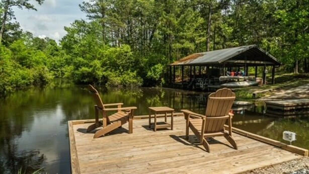 Een houten steiger met Adirondack-stoelen en een kleine tafel, met uitzicht op een rustig meer met een overdekte zithoek op de achtergrond.