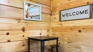 Een houten hutje van binnen, met een 'WELCOME' bord, een kleine tafel en twee krukken.