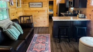Een gezellig chaletinterieur met houten details, een verwelkomingsbord, een keuken met een bar en een zithoek met een bank en voetenbank.