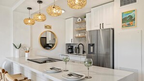 A modern kitchen with a central island, white cabinetry, a stainless steel refrigerator, and hanging pendant lights. - SUGAR BEACH BLISS #2- BRAND NEW Designer Luxury-Ocean/Volcano Views 5 STAR Owner (Kihei)