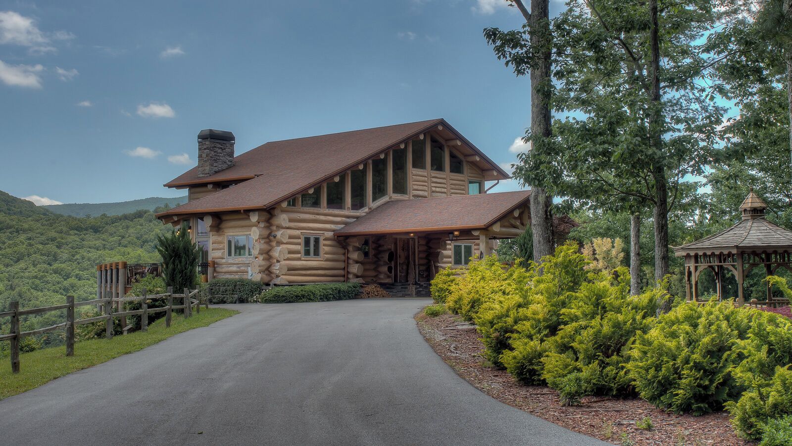 A log cabin with a stone chimney, a porch, and a gazebo in a forested area.