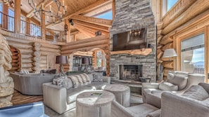 L’intérieur d’une cabane en rondins est agrémenté d’une cheminée en pierre, d’un grand téléviseur à écran plat fixé au-dessus, et d’un coin salon confortable avec des canapés et des poufs.