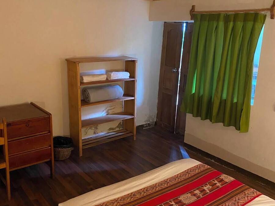 Quarto com cama e cômoda de madeira, uma estante e uma janela com cortinas verdes.