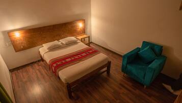 Un dormitorio con cabecera de madera, cama con sábanas blancas y una manta roja estampada, un sillón color turquesa y una mesita de noche de madera.