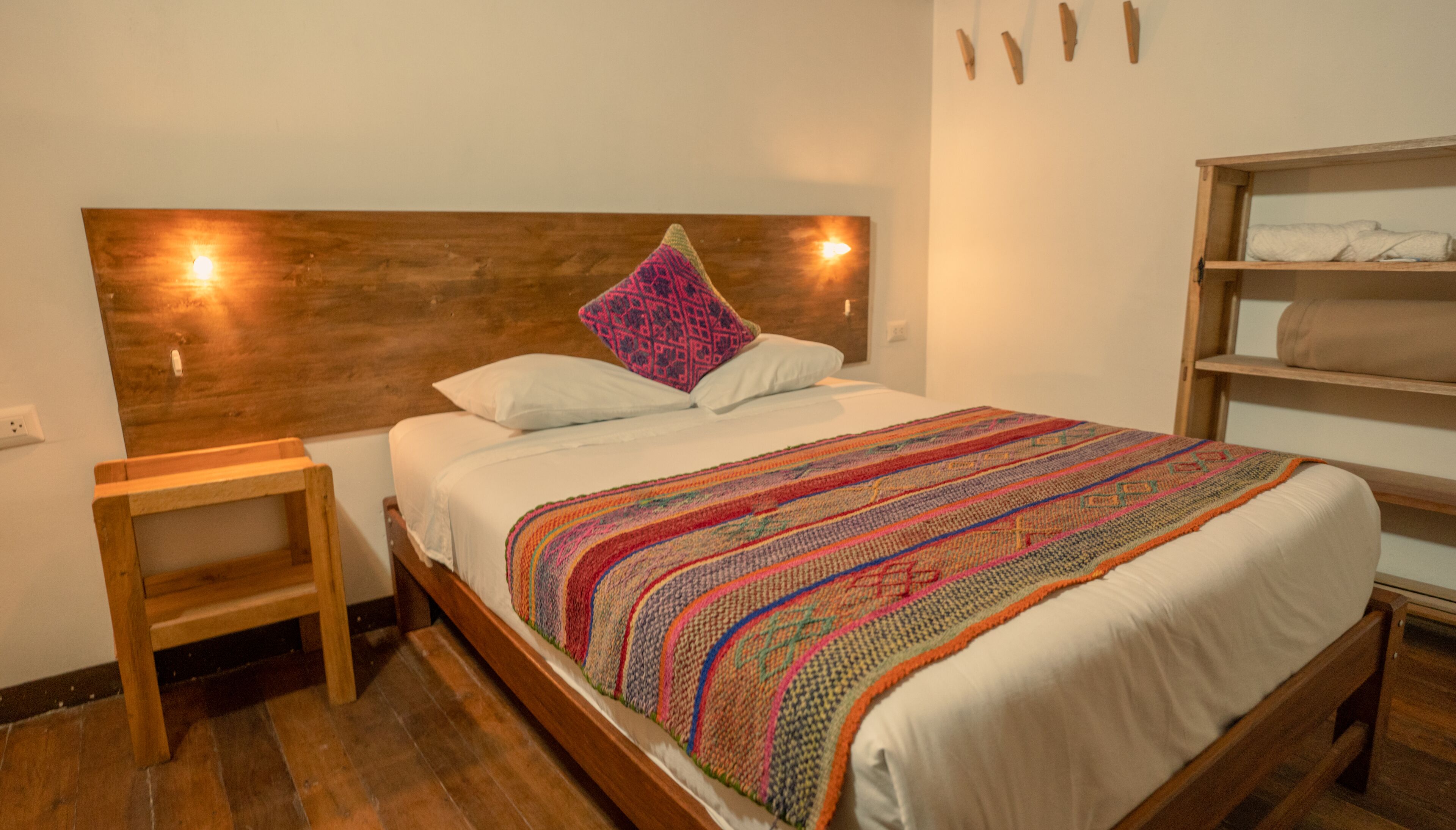 Un lit bien fait, avec une couverture colorée, une tête de lit en bois, une petite table de chevet en bois et une étagère où sont pliées des serviettes.