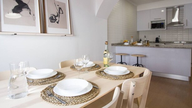 A dining area with a table set for four, framed artwork on the wall, and a kitchen in the background. - Comfortable getaway in Medieval Castro Marim by Lisbeyond (Castro Marim)