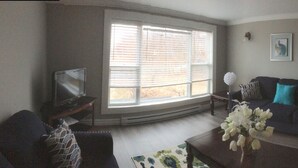 Ein Wohnzimmer mit einem dunkelgrauen Sofa, einem kleinen Tisch mit Blumen und einem Fenster mit Jalousien. An der Wand hängen zwei gerahmte Bilder und ein dekorativer Spiegel.