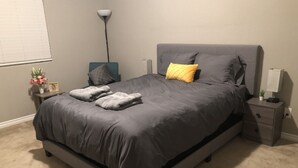 Quarto moderno com uma cama cinza, uma mesa de cabeceira com abajur e uma mesinha de apoio com uma planta.