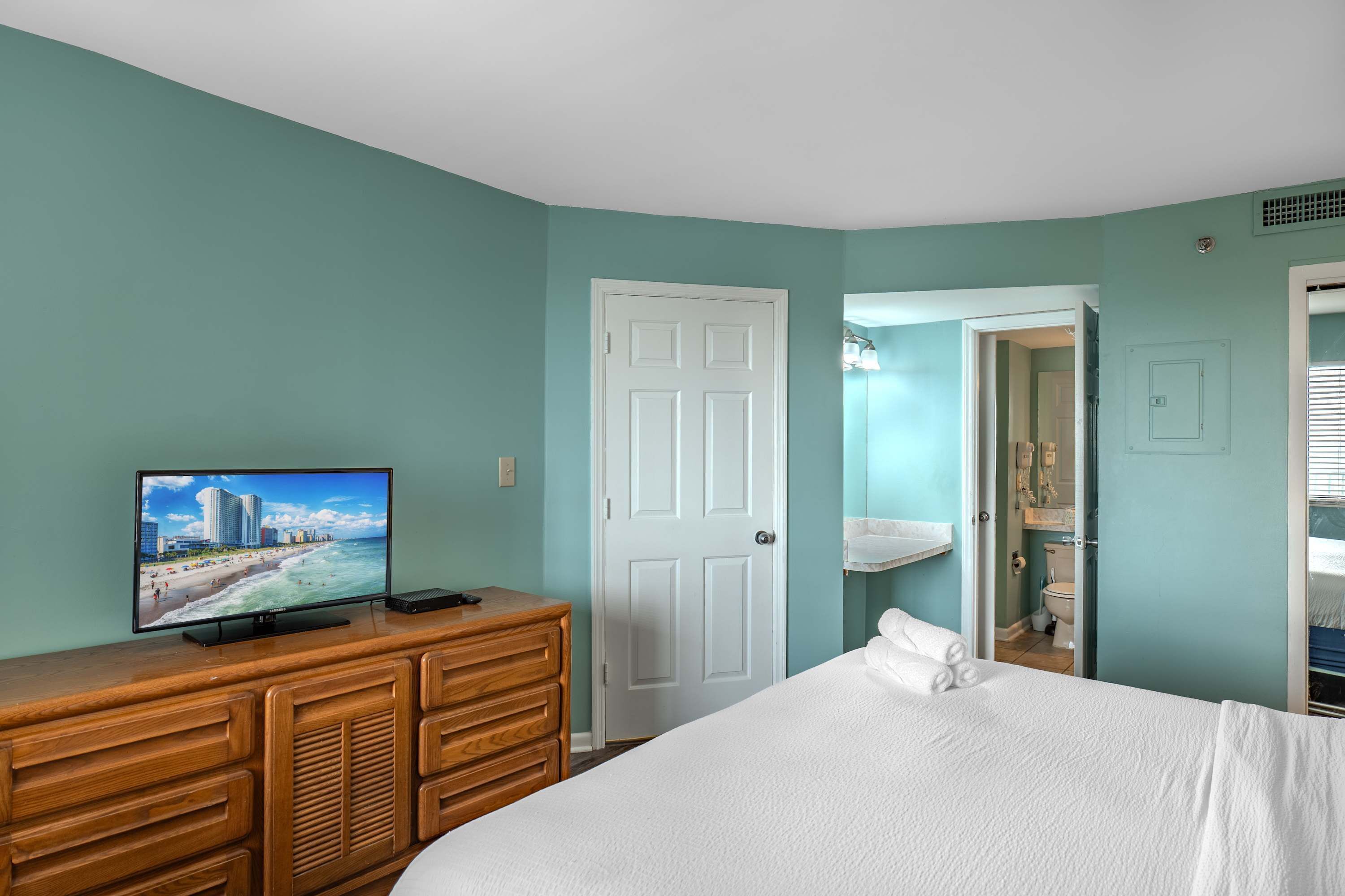 A bedroom with a bed, dresser, television, and a bathroom visible through an open door.