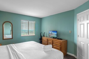 A bedroom with a bed, a dresser, a television, a window with blinds, and a door.