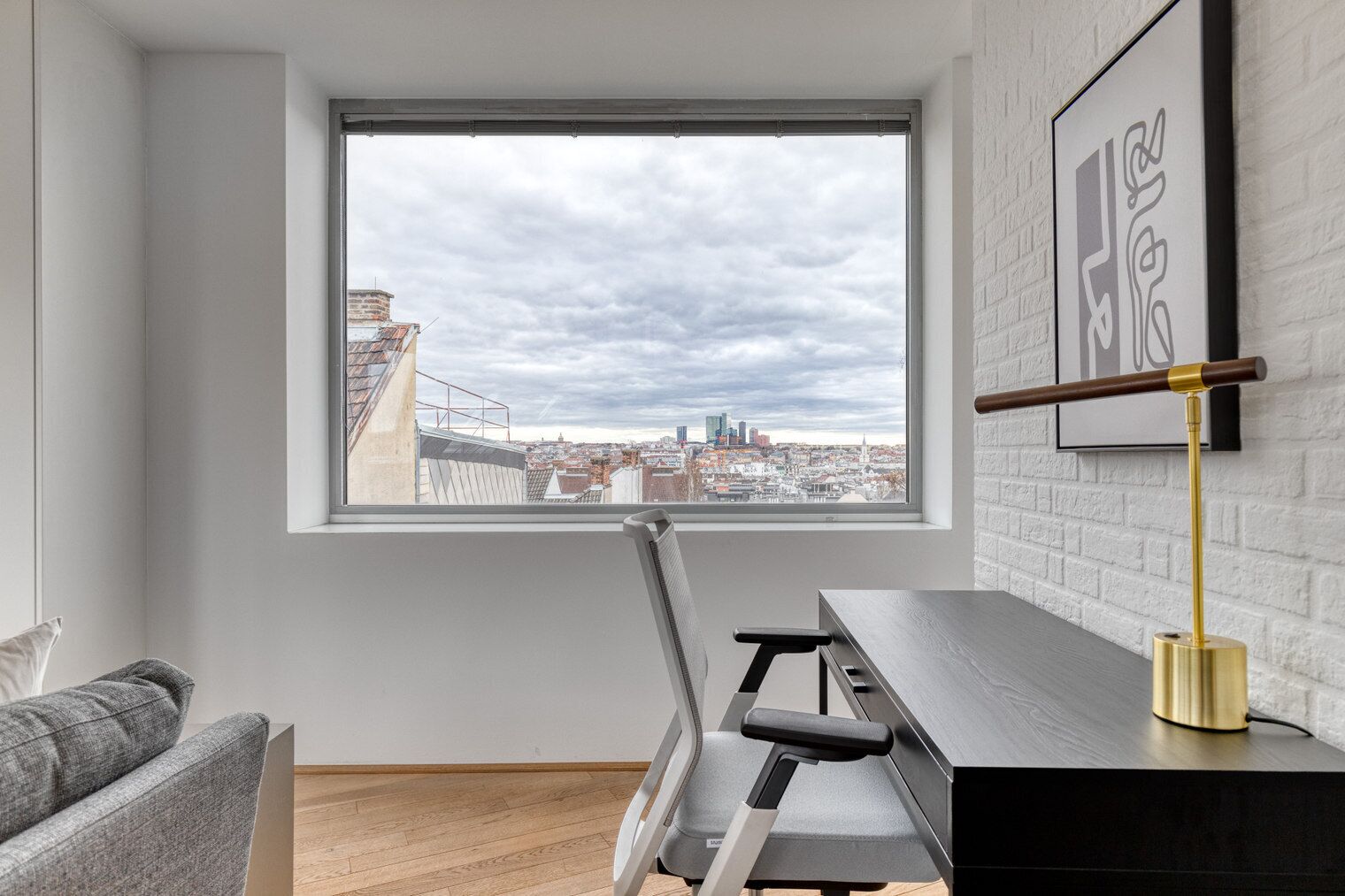 A room with a view of a cityscape through a large window, a treadmill, a desk with a lamp, and a grey sofa.