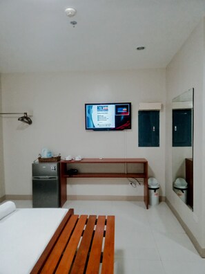 A hotel room with a bed, a wooden bench, a TV mounted on the wall, a small desk, and a trash bin. - Grace & RB Hotel (Kalibo)