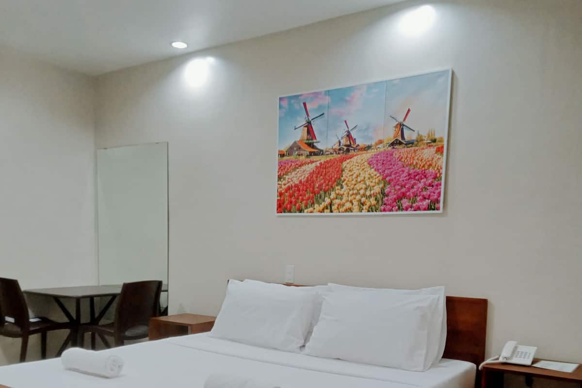 Habitación de hotel con una cama de madera, ropa de cama blanca, un cuadro con molinos de viento y flores en la pared, un escritorio con sillas y una mesita con un teléfono.