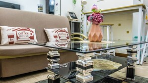 A living room with a glass coffee table, a beige sofa with coffee-themed cushions, and a vase with flowers on a glass table. - Stylish 1BR Retreat at Trees Residences — Fast WiFi, Smart TV, Near SM Fairview (Quezon City)