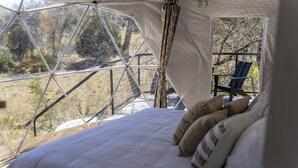 A bed with pillows in a tent-like structure with a view of trees and a chair outside.