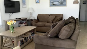 A living room with a brown sofa, a TV, a table with books, and a vase of flowers.