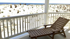 A wooden lounge chair on a balcony overlooking a beach with sparse vegetation and a clear sky.