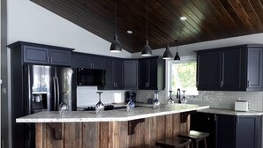 A modern kitchen with dark cabinets, a large island with stools, stainless steel appliances, and pendant lighting. - Where Friends and Family make Memories. (South River)
