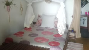 Una habitación con una cama bajo un dosel blanco, un banco de madera, una mesita y una planta en maceta.