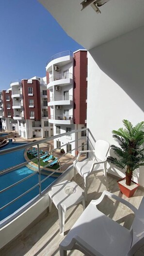 A balcony with a pool, white lounge chairs, and a potted plant. - 1 Bed Apartment - Balcony, Pool, Parking (Hurghada)