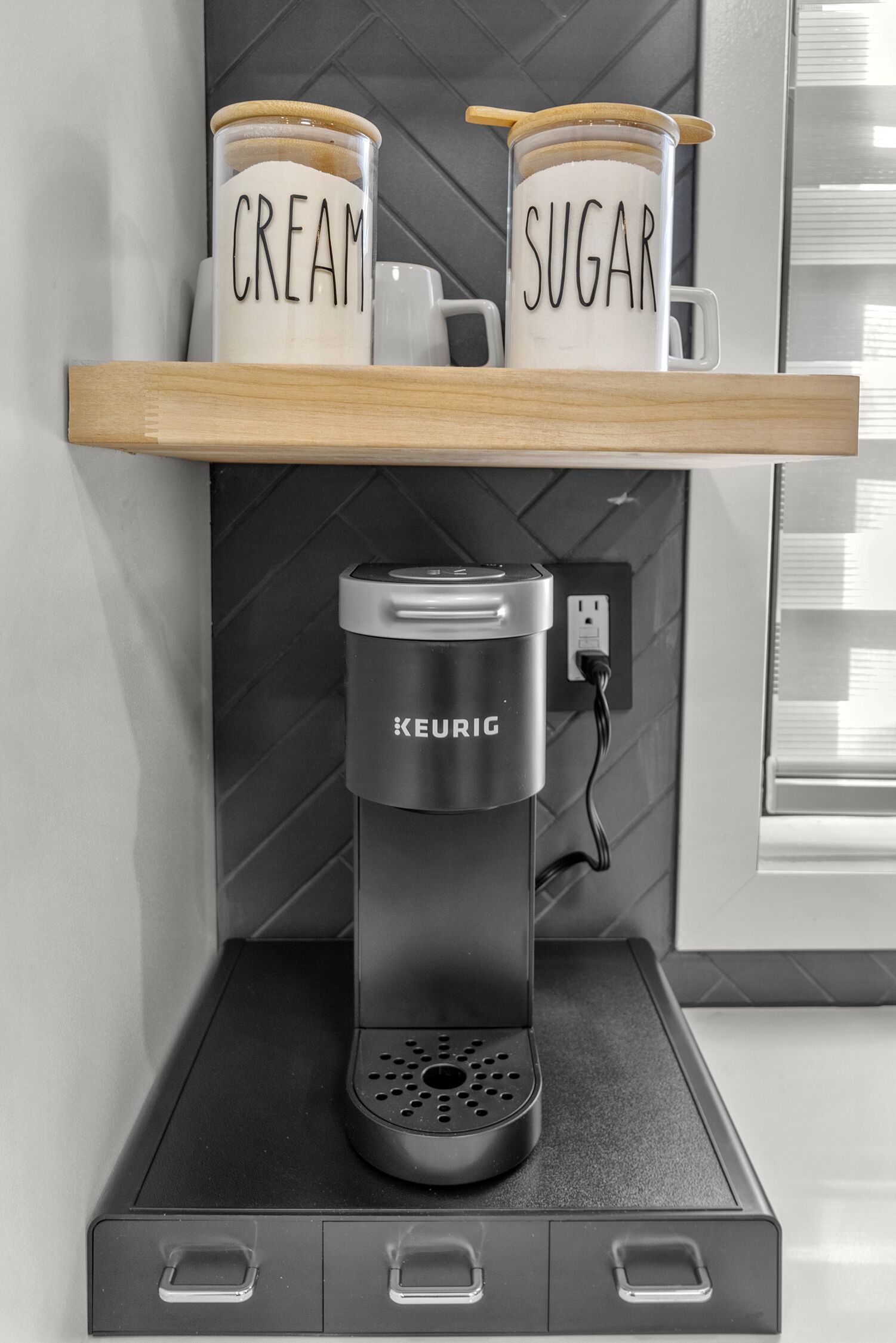 A Keurig coffee maker on a black countertop with a shelf above it holding two white mugs labeled "CREAM" and "SUGAR".