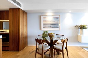 A dining area with a round glass table, wooden chairs, a framed picture on the wall, and a kitchen in the background.