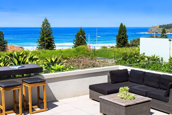 An outdoor seating area with a view of the ocean, a sofa, two stools, and a table with a vase.