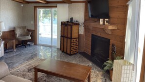 A spacious living room with a fireplace, wooden beams, a television mounted on the wall, and a large window. - Waterfront Bungalow nightly panoramic view of our illuminated covered Bridge (Galloway)