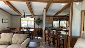 A spacious living area with a wooden ceiling, a dining table with chairs, a sofa, and a kitchen with wooden cabinets and a window. - Waterfront Bungalow nightly panoramic view of our illuminated covered Bridge (Galloway)