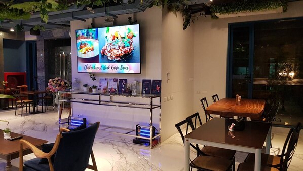 A restaurant interior with a TV displaying food images, a counter with a menu, and a dining area with tables and chairs. - BlueTel Re'sidence Bangkok IMPACT (Pak Kret)