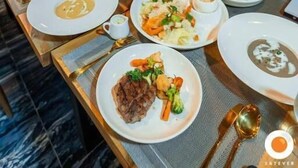 A restaurant with a variety of dishes on the table, a view of the city at night through large windows, and hanging pendant lights. - BlueTel Re'sidence Bangkok IMPACT  (Pak Kret)