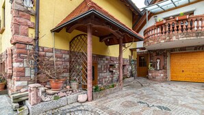 Sisäpiha, jossa on mukulakivilaatoitus, tiiliseinä, puinen pergola ja keltainen rakennus, jossa on puuovi.