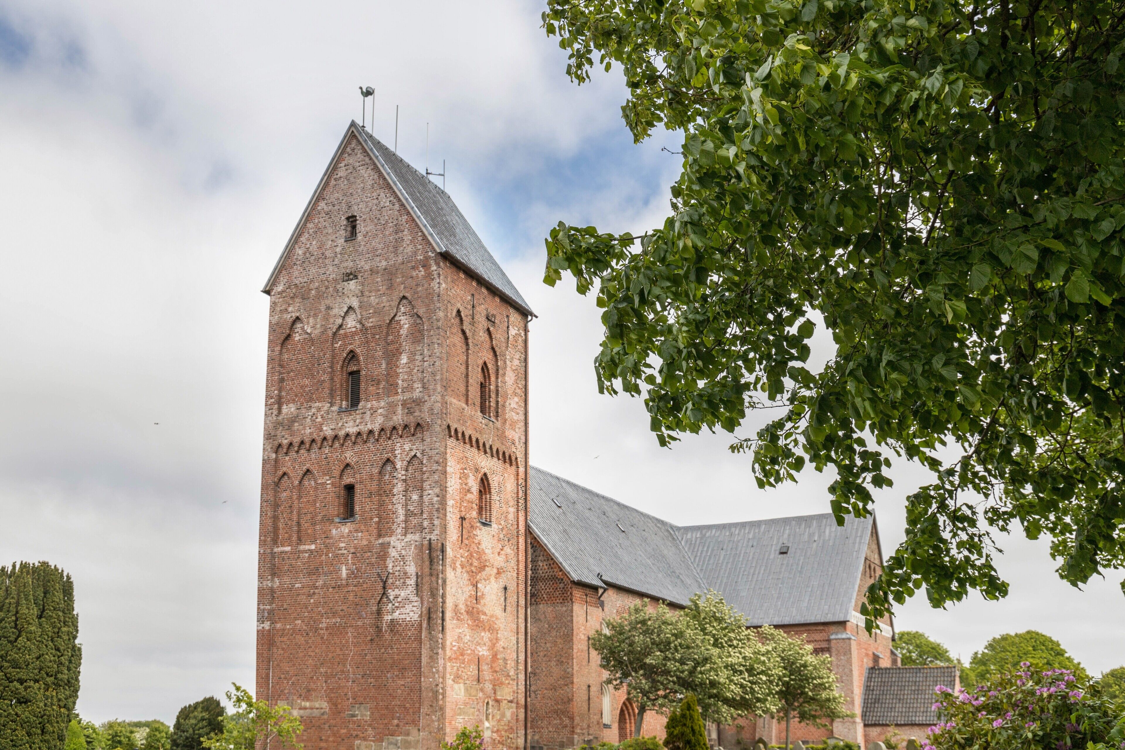 En historisk byggnad i tegel med ett markant torn, omgiven av träd och en klar himmel.