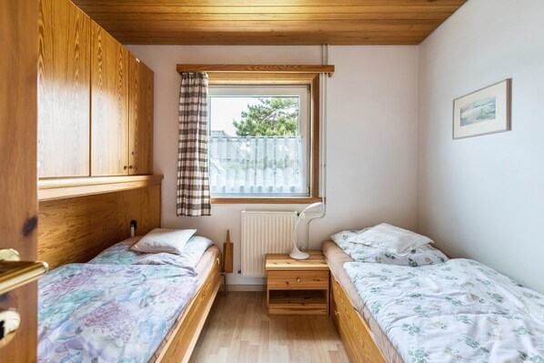 A small, well-lit bedroom with two beds, a window with a view, a radiator, and wooden furniture.