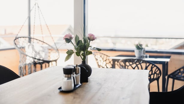 Une table en bois ornée d’un vase de fleurs roses et de salières et de poivrières, un fauteuil suspendu et une vue sur la plage.