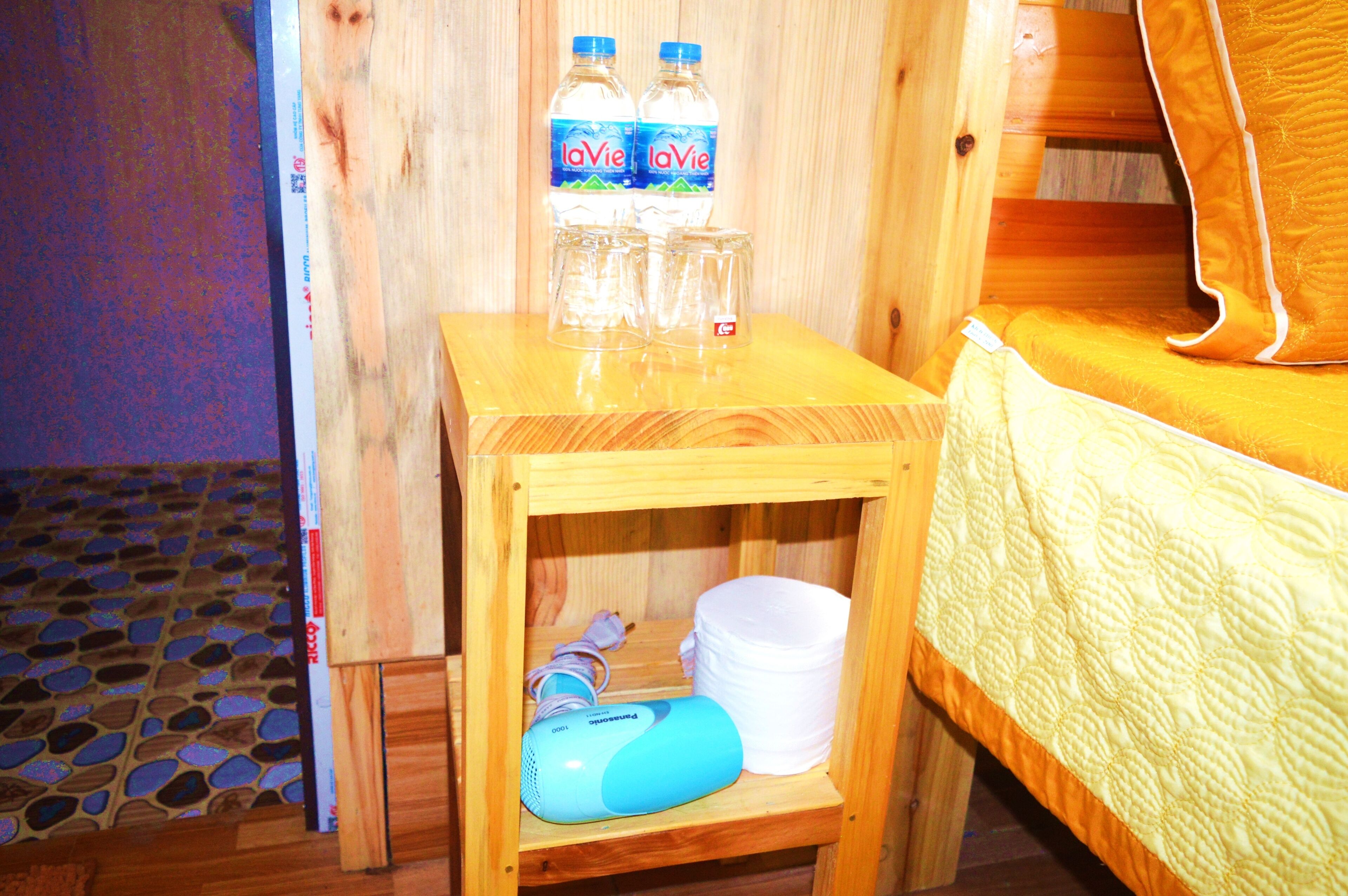 Un tavolo in legno con bottiglie d'acqua, bicchieri e un dispositivo blu in una stanza con un letto e un pavimento decorato.