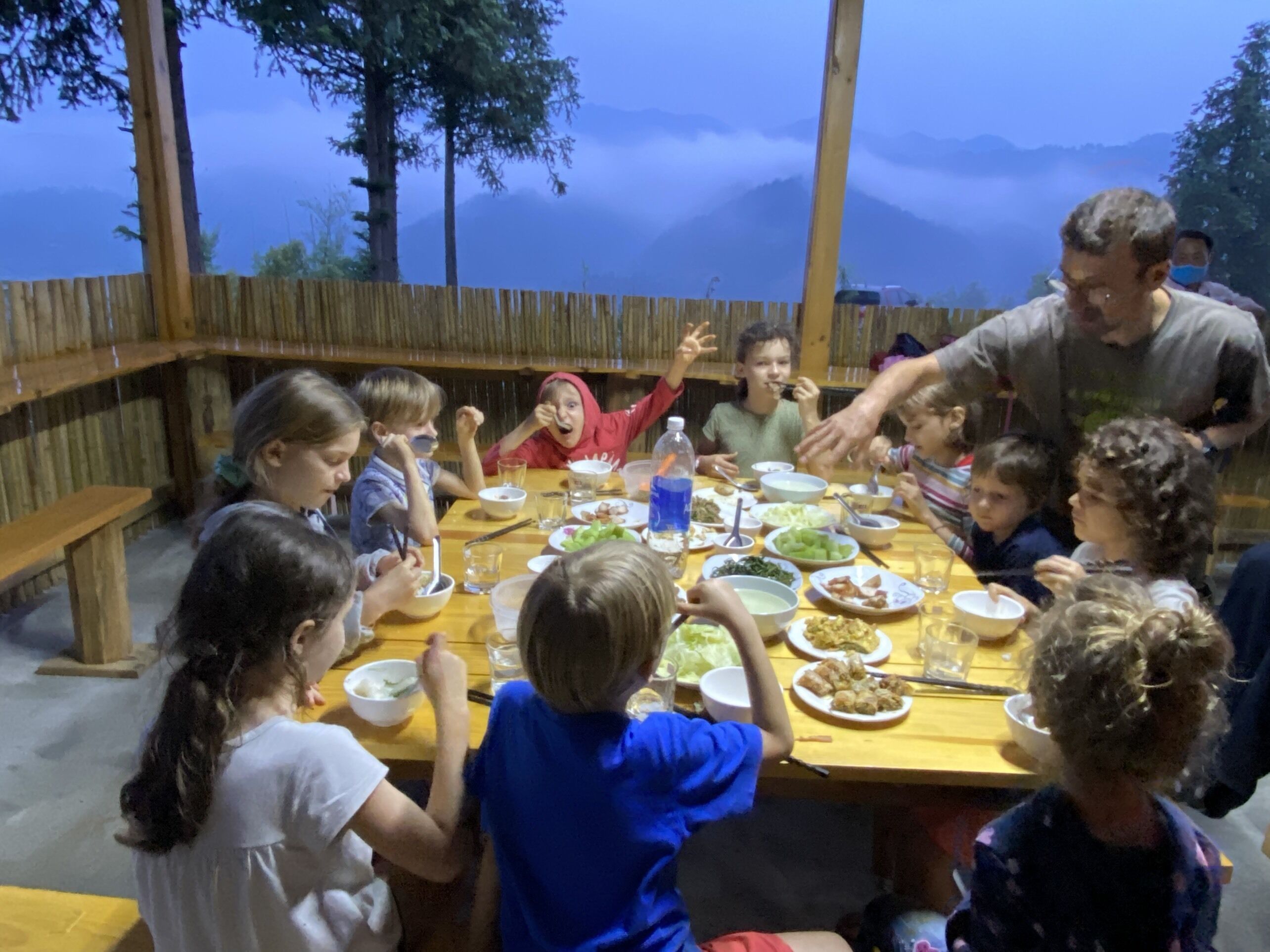 Um grupo de pessoas jantando em uma mesa de madeira ao ar livre, com montanhas ao fundo.