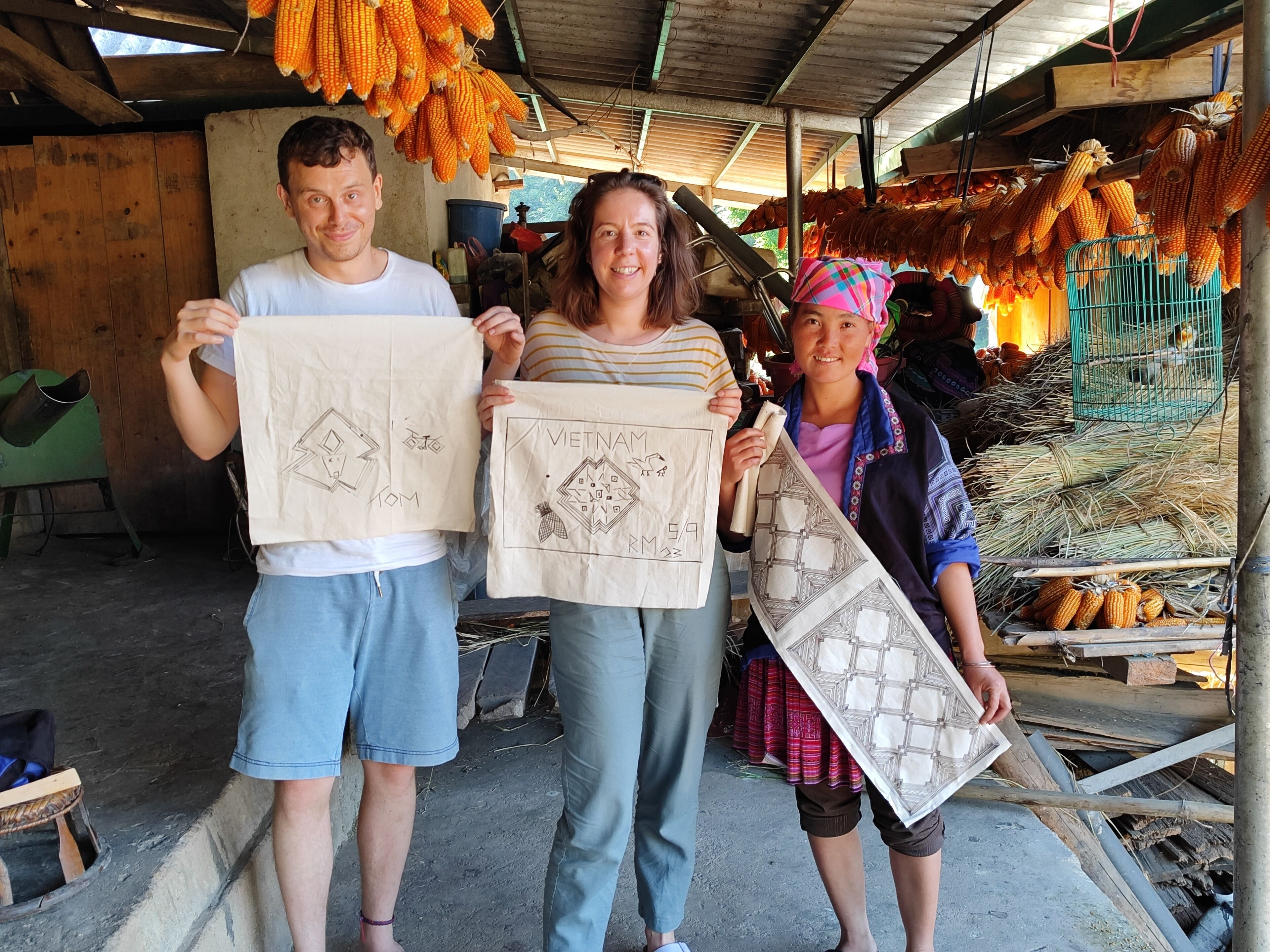 Three people holding bags with Vietnamese designs, standing in a market with corn on display.
